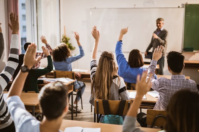students raising hands; POV back of classroom